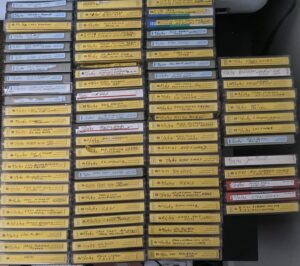 Photograph of dozens of cassette tape cases, each with hand-written labels indicating air date and topic of that episode.